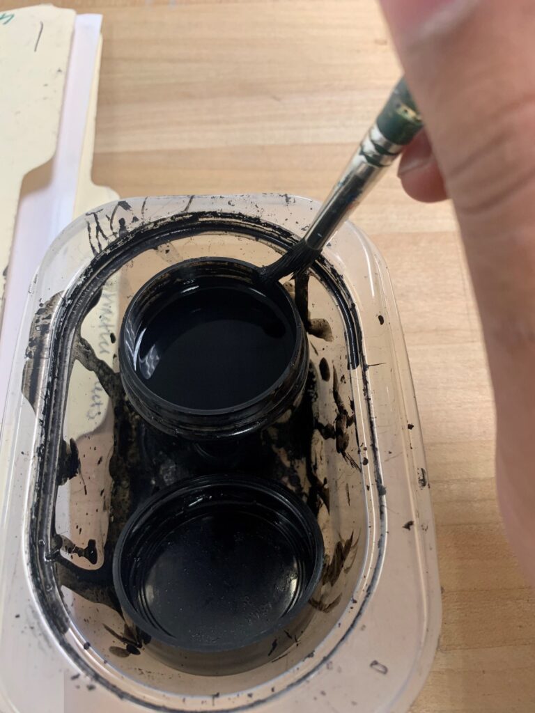 A rounded brush just outside of a small container of black ink, being rubbed on the edge of a clear plastic container.