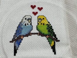 A completed cross stitch project depicting two birds (one blue and white, one green) on a branch with three red hearts hovering above between them.