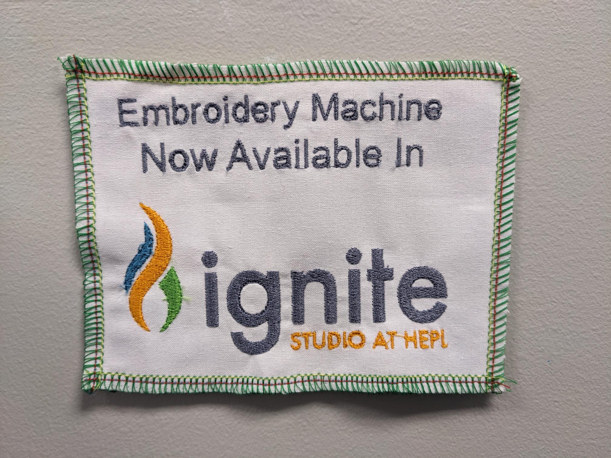 An embroidered design that reads "Embroidery Machine Now Available in Ignite Studio at HEPL" with the Ignite flame logo.