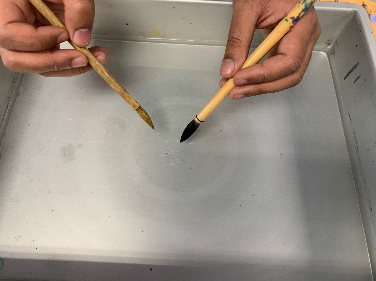two hands holding two calligraphy brushes dipping one into the water resulting in a round ink shape