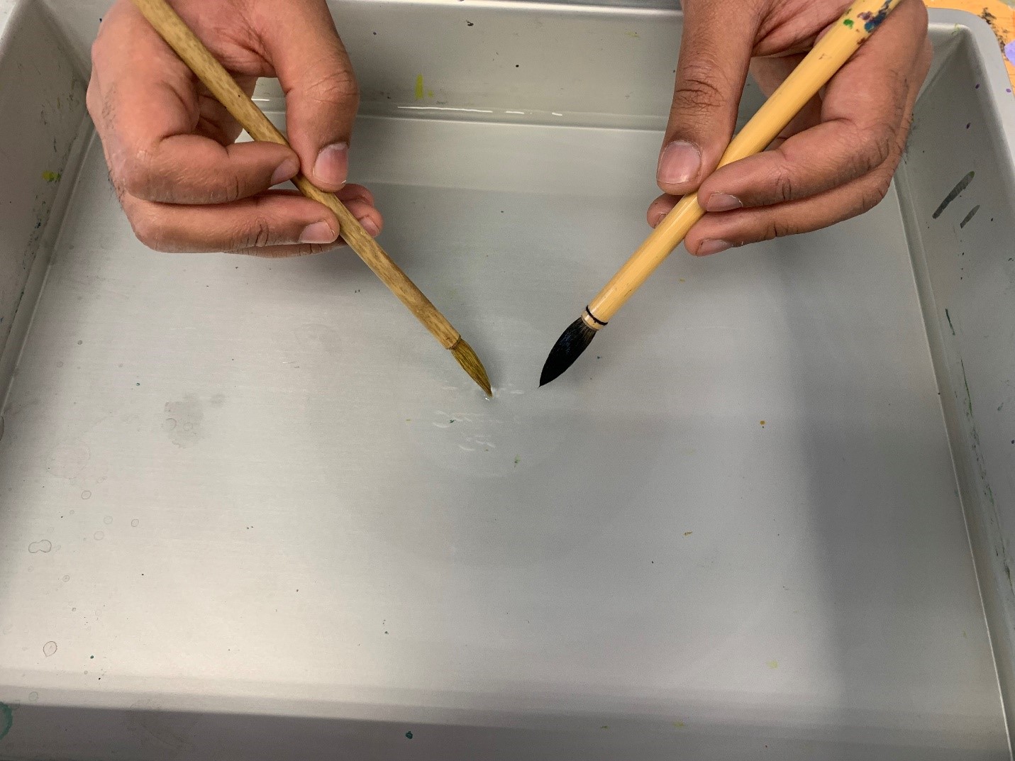 two hands holding two calligraphy brushes dipping one into the water resulting in another round ink shape inside the first