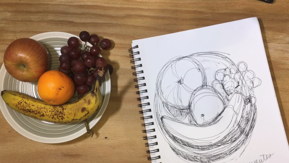 An overhead view of a bowl of fruit next to a sketchpad depicting a contour line drawing of the fruit bowl.